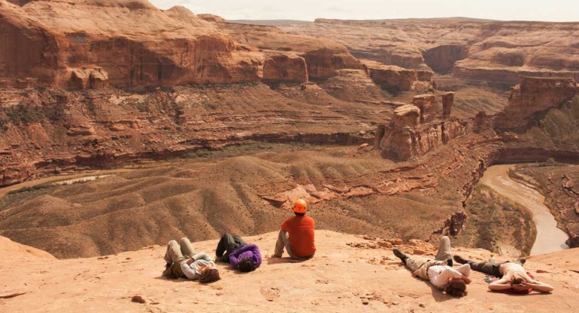 People rest atop a rocky overlook, with red canyon walls and a river below. 
